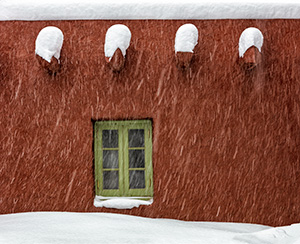 Snow piles on top of wooden support beams on a terra cotta-colored wall above a light green wooden window frame. By Claire Curran