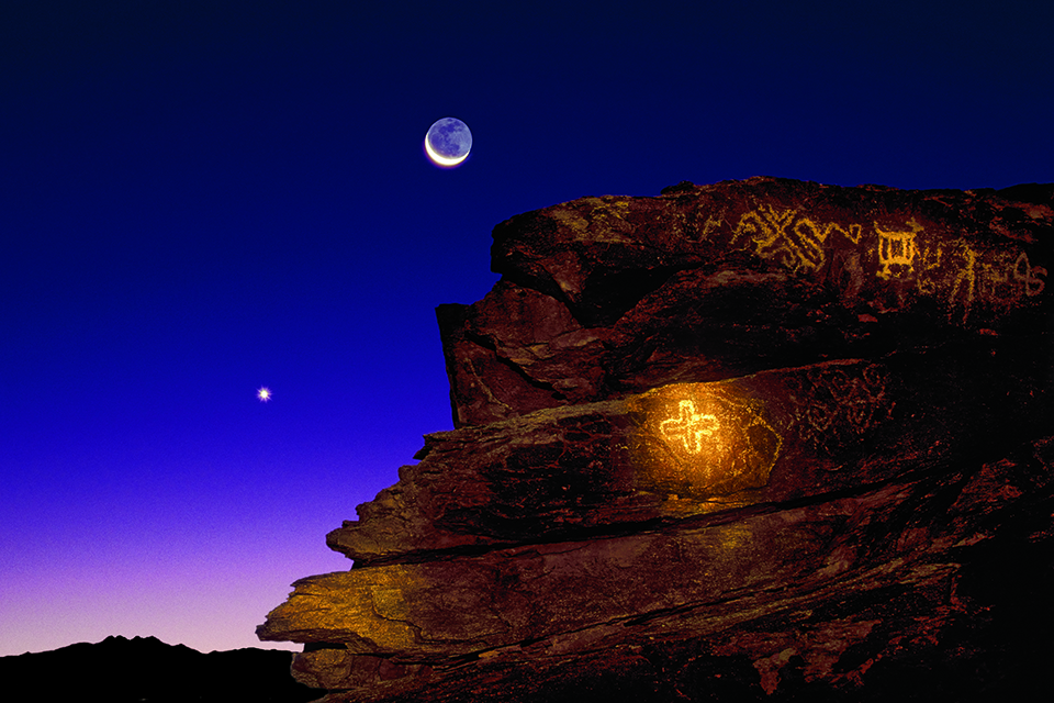 Venus and a crescent moon set in a sky barely touched by dawn, while a Hohokam cross — a symbol for Venus — illuminated with a flashlight, brightens a rock outcropping at South Mountain Park in Phoenix. By Frank Zullo