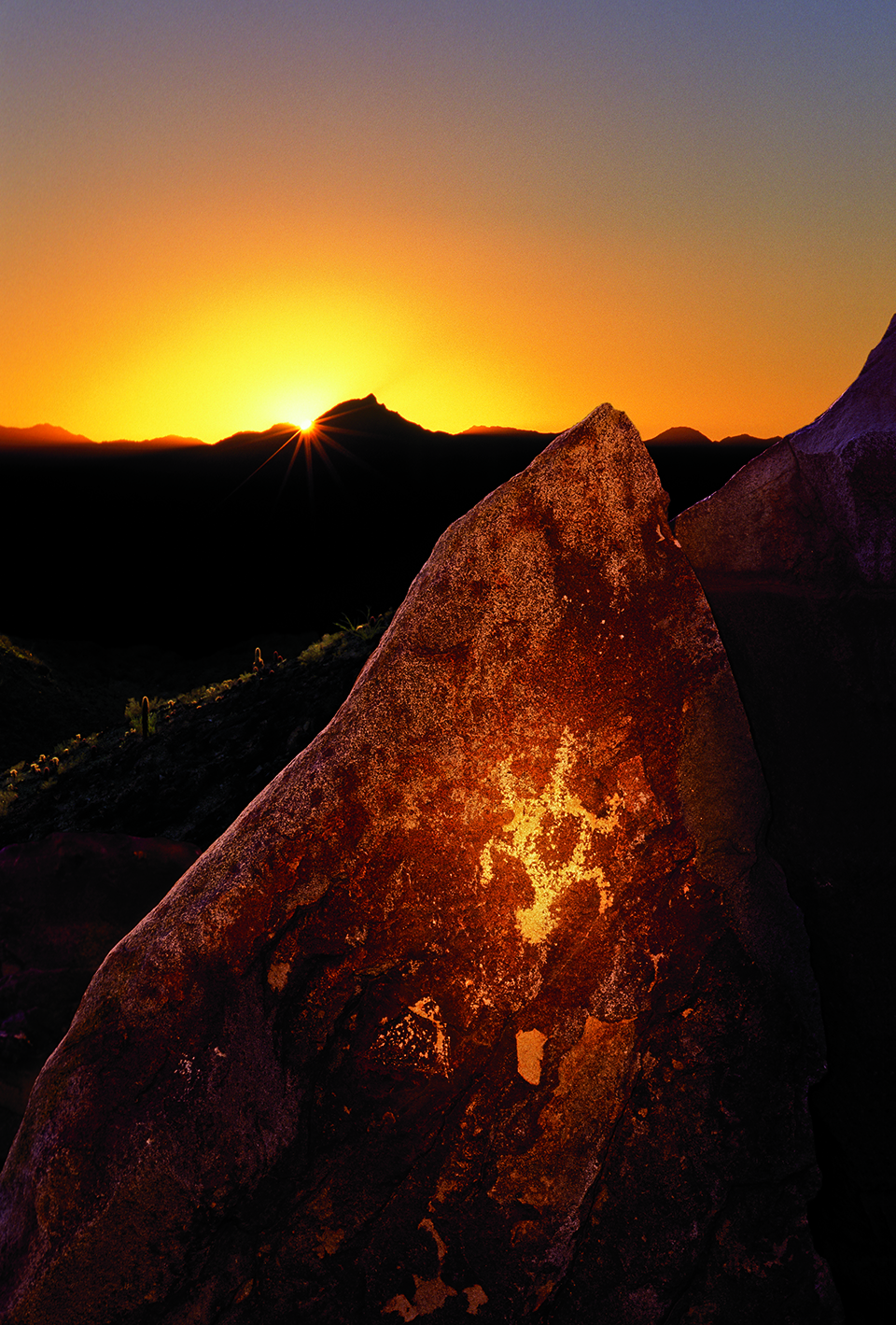 On the winter solstice, the last rays of the setting sun stream through a notch in the Sierra Estrella Mountains, as viewed from a prehistoric Hohokam sun-watching shrine. The highlighted shrine, marked by a lone lizard petroglyph, appears on a monolith at South Mountain Park in Phoenix. By Frank Zullo