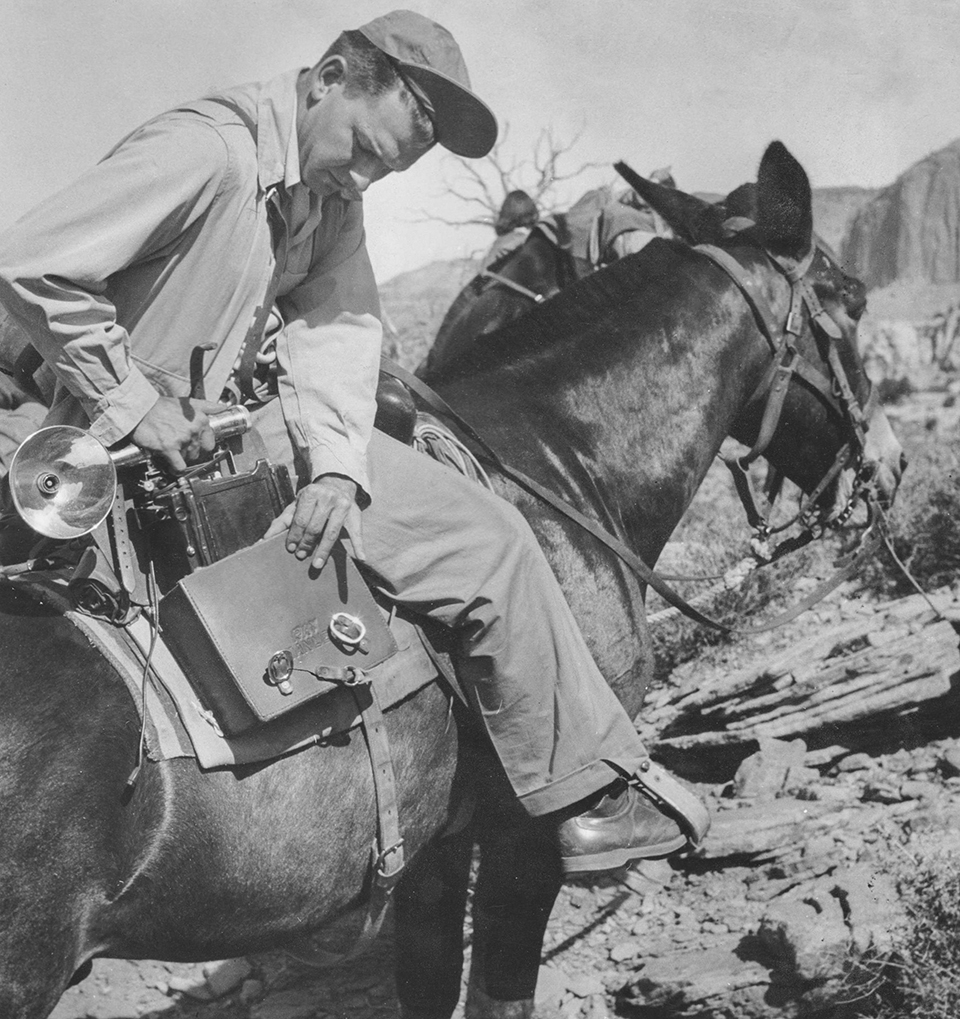 BW photo of Ray Manley lifting a 4x5 camera out of a bag while mounted on a mule in the desert. | COURTESY OF CAROLYN MANLEY ROBINSON