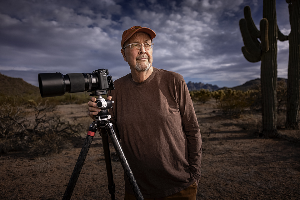 Jack Dykinga poses under a cloudy sky in the desert with a long-lensed camera mounted on a tripod. | JOEL GRIMES 