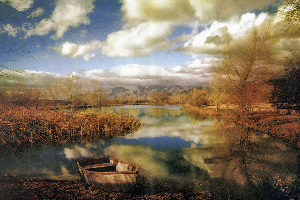 This painterly photograph by Esther Henderson shows autumn foliage and cloudy blue skies reflected in a lake with mountains in the background.