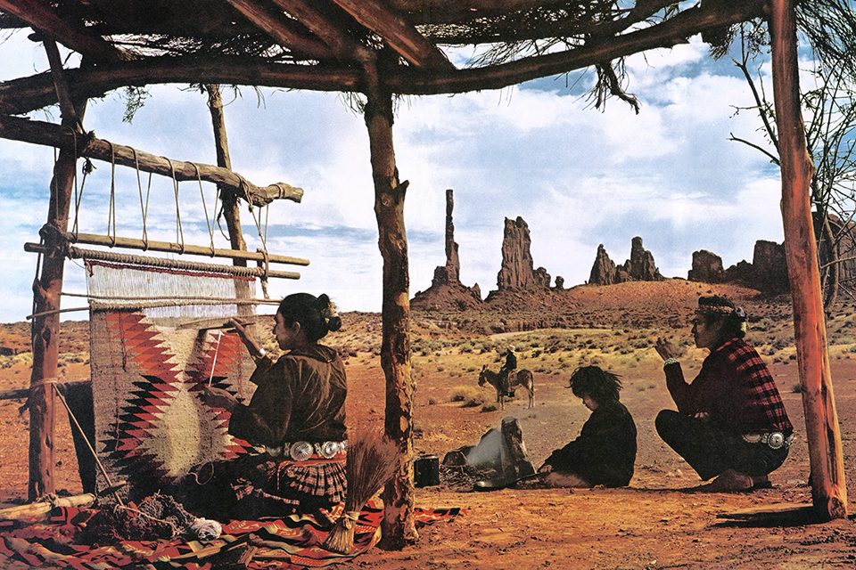 Josef Muench photo of Navajo woman weaving a rug with her family nearby with Yei Bichei rocks of Monument Valley as a backdrop.