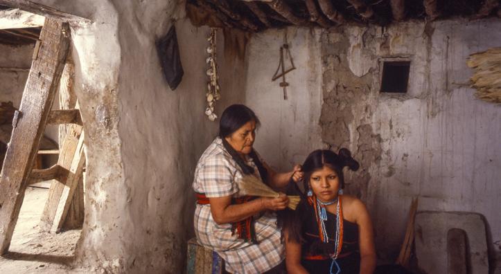 One indigenous woman sits on a crate, fixing another indigenous woman's hair.